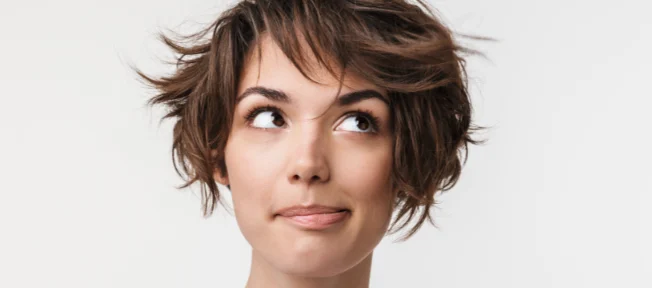 A woman with fair skin and choppy brown hair looks up to the left in front of a white background.