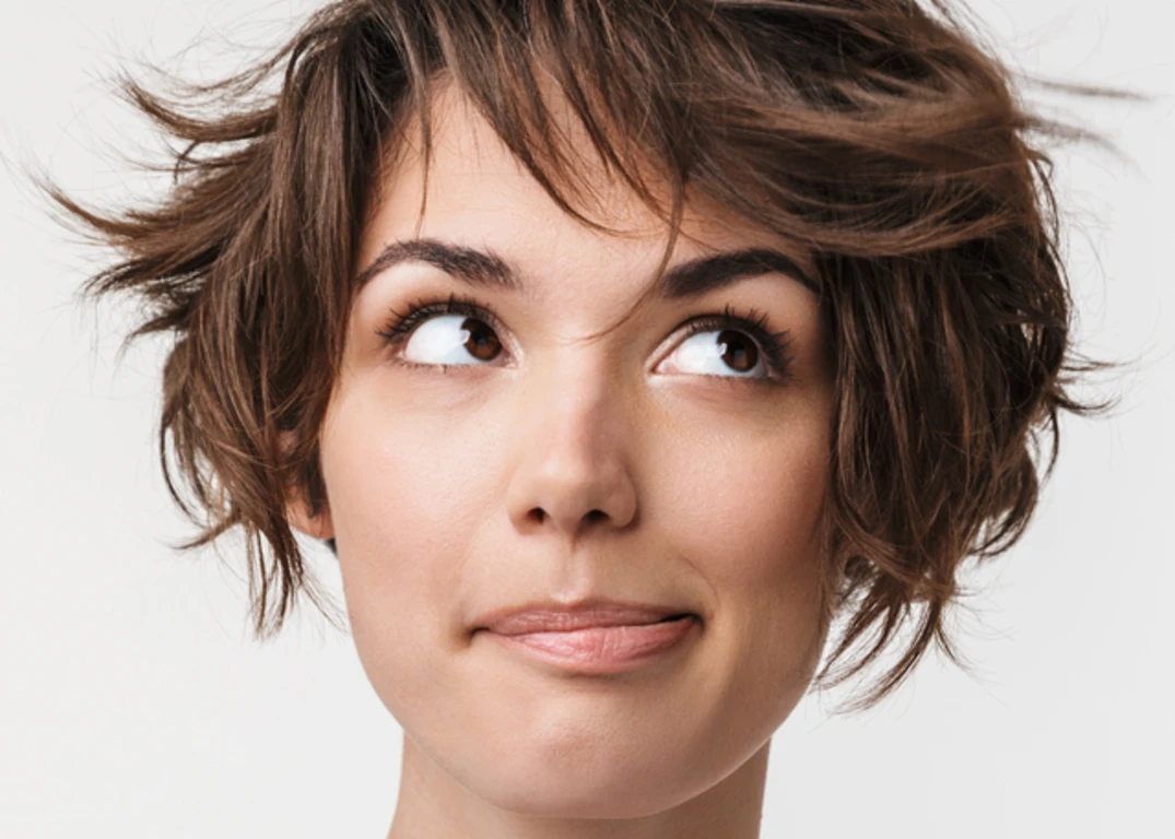 A woman with fair skin and choppy brown hair looks up to the left in front of a white background.