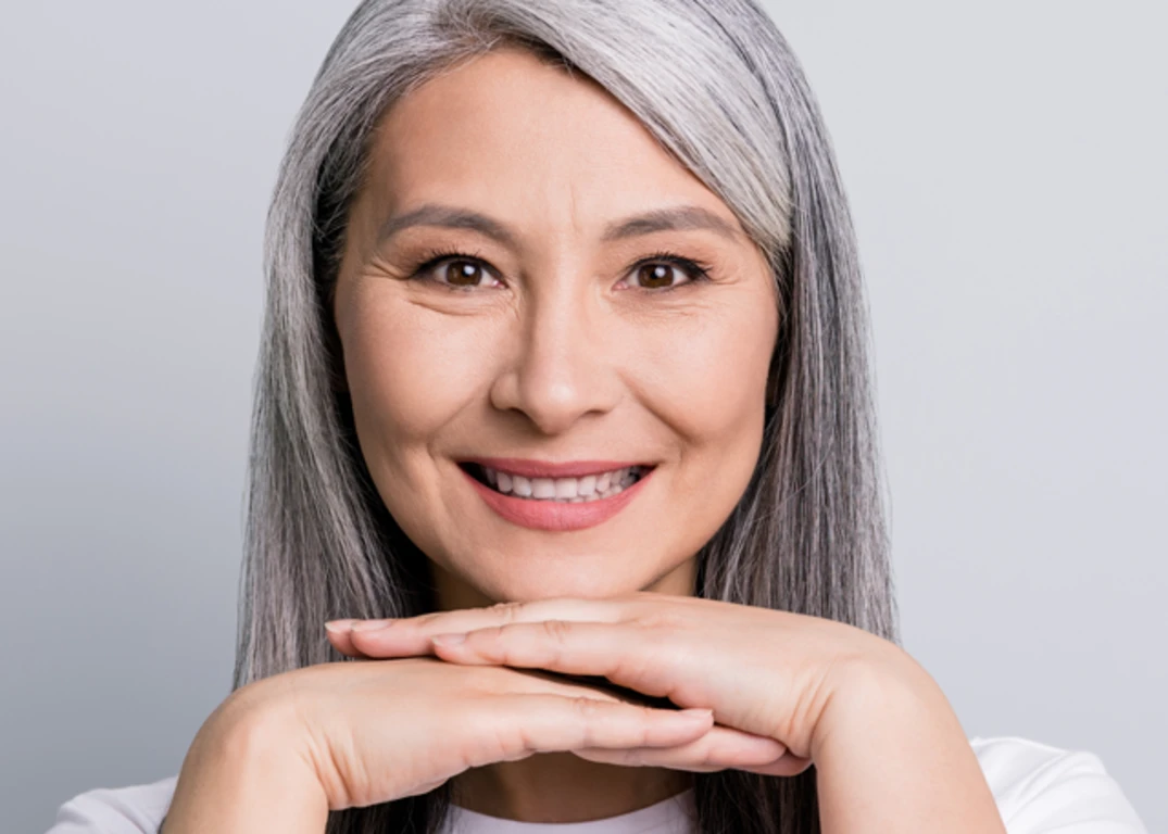 A woman with long gray hair smiles facing forward with her hands below her chin in front of a white background.