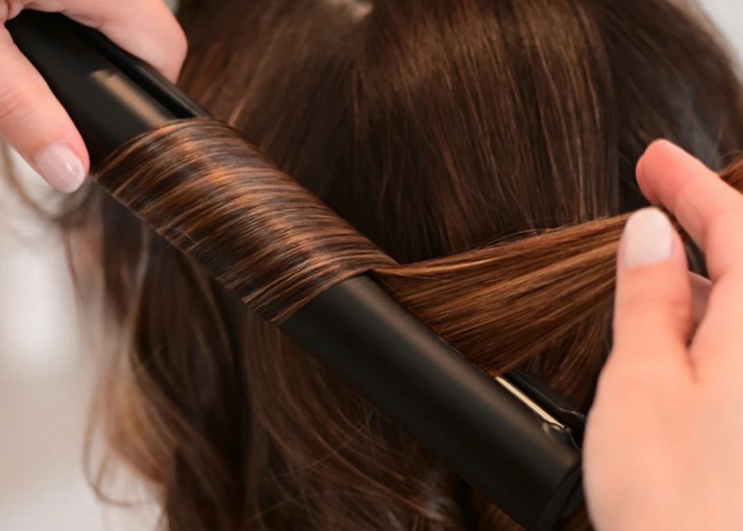 A stylist uses a black flat iron to curl a woman’s brown hair.