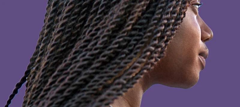 A dark-skinned woman with hair in a senegalese twist looks to the right in front of a purple background. 