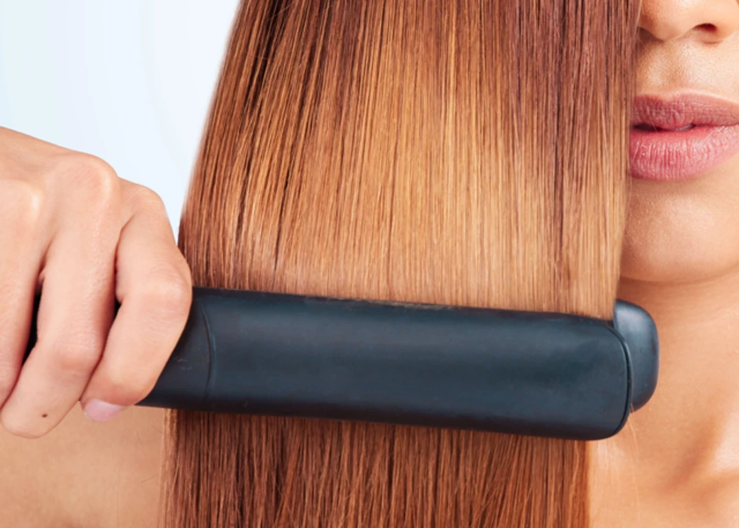 Woman with tan skin and yellow brown hair uses flat iron to straighten her hair in front of a grey background