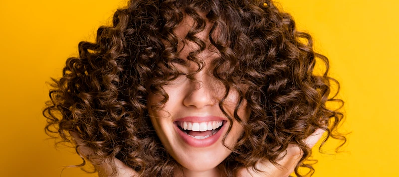 A woman with curly brown hair holds it up with flat hands in front of a yellow background.