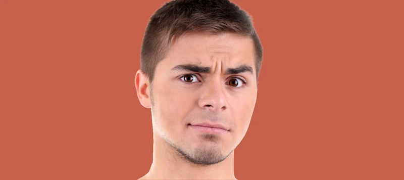 a man with fair skin and short brown hair looks inquisitive in front of an orange background.