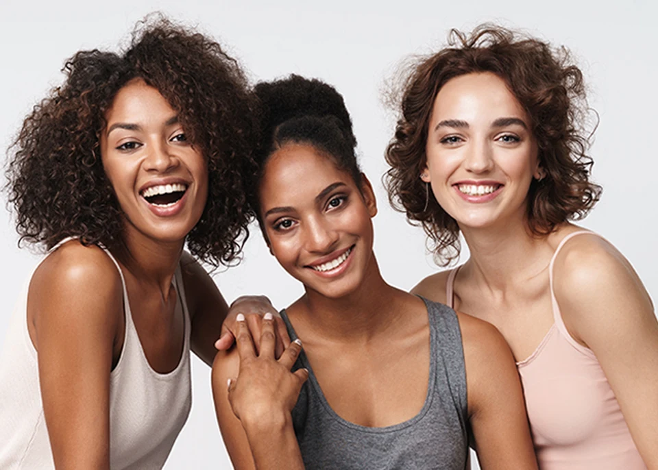 Portrait of three gorgeous multiracial women standing together with a combination of curly and wavy hair and smiling at camera isolated over white background