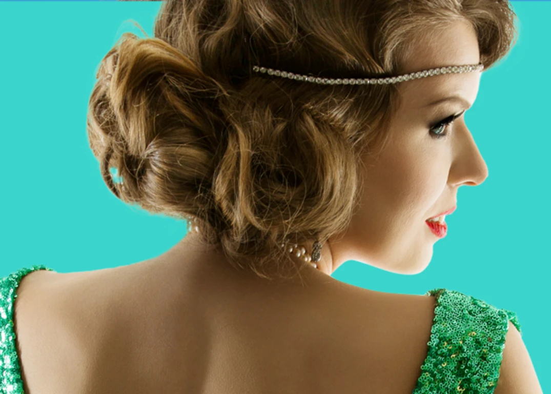 A woman with fair skin and a 1920’s hairstyle faces away and looks to the right wearing a green sparkling dress in front of a turquoise background.
