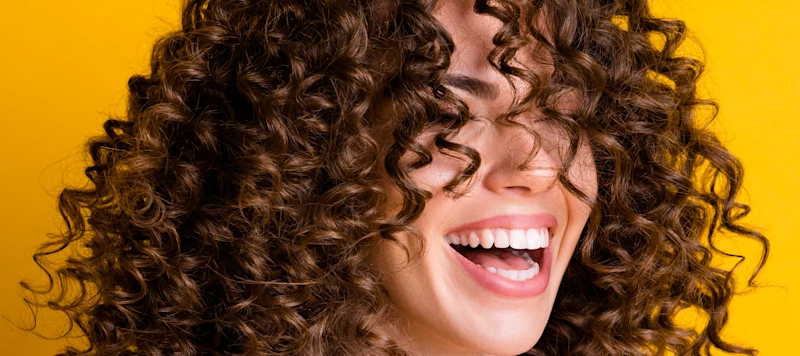 a woman with curly brown hair smiles with her eyes closed in front of a yellow background.