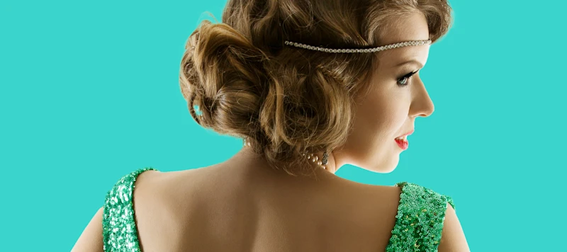 A woman with fair skin and a 1920’s hairstyle faces away and looks to the right wearing a green sparkling dress in front of a turquoise background.