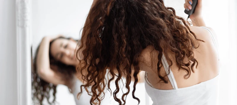 Young brunette girl female woman adult in grey shorts and white top drying her curly hair with blow dryer after shower in front of the mirror.