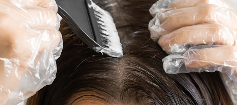 A woman uses gloves and a brush to apply dye to her dark brown hair at the roots.