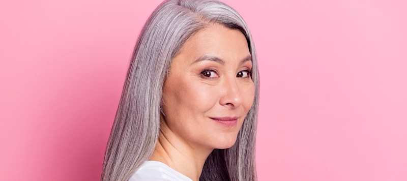 a woman with a round face and long gray hair turns her head to the right in front of a pink background.