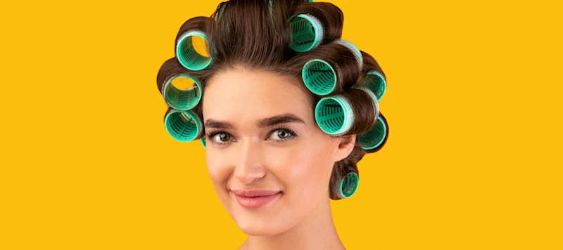 A woman with green heated rollers in her brown hair looks forward in front of a yellow background.