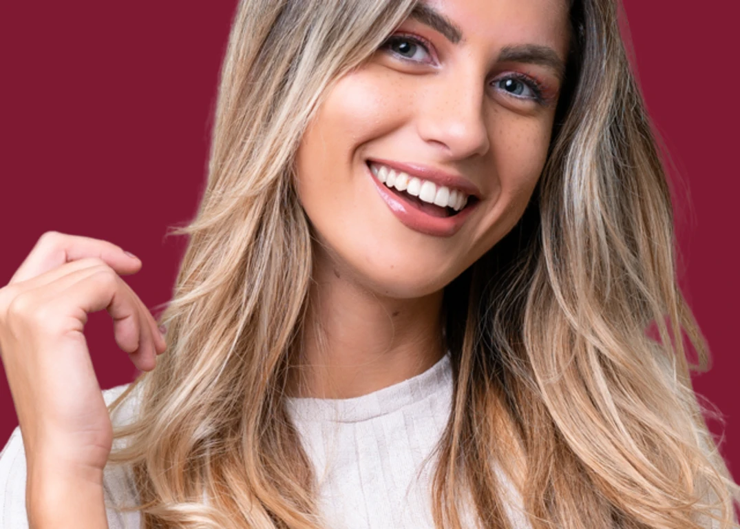 A woman with fair skin and demi permanent blonde hair smiles facing forward in front of a maroon background.