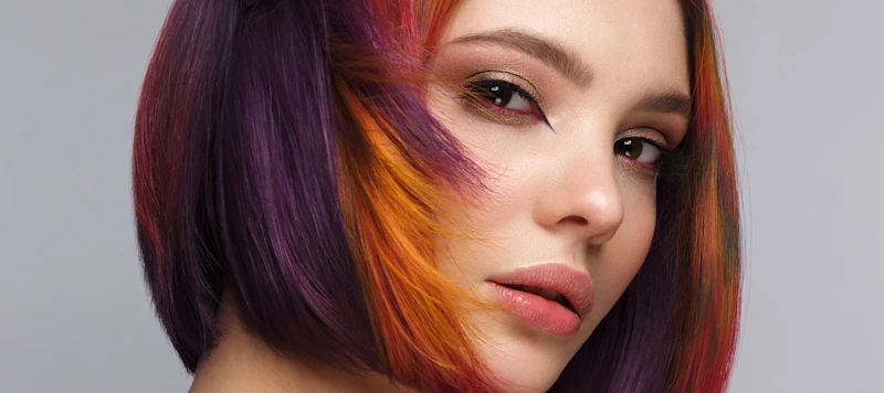 A woman with fair skin and demi permanent hair dye for short red, purple and orange hair looks forward in front of a gray background.