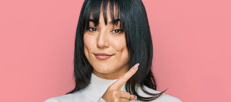 A woman with tan skin and medium length black hair and bangs grins facing forward in front of a pink background.