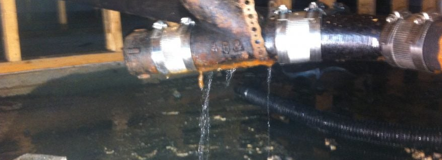 This image shows a rusted metal pipe with water leaking from a crack, despite being secured with metal clamps, indicating plumbing damage and the need for repair.