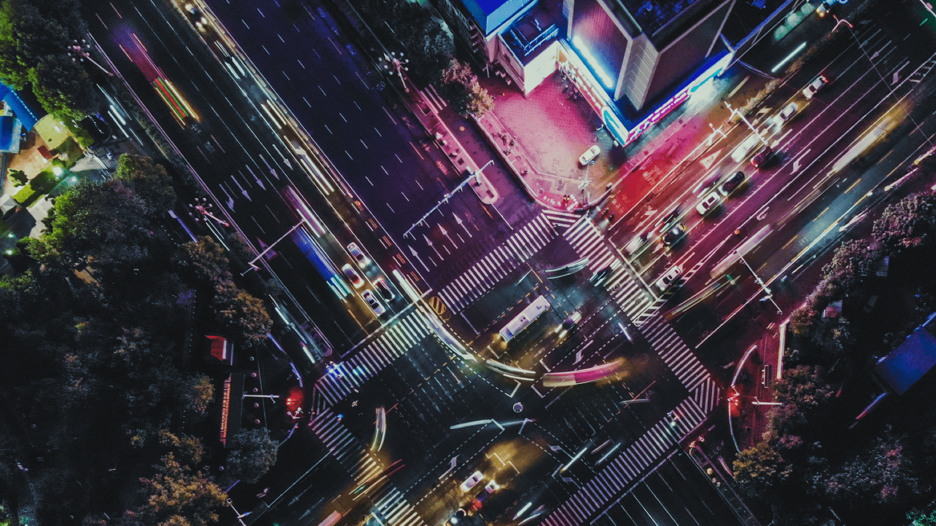 Aerial view of a busy city intersection at night with glowing lights, outdoor digital displays and moving vehicles.