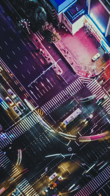 Aerial view of a busy city intersection at night with glowing lights, outdoor digital displays and moving vehicles. 