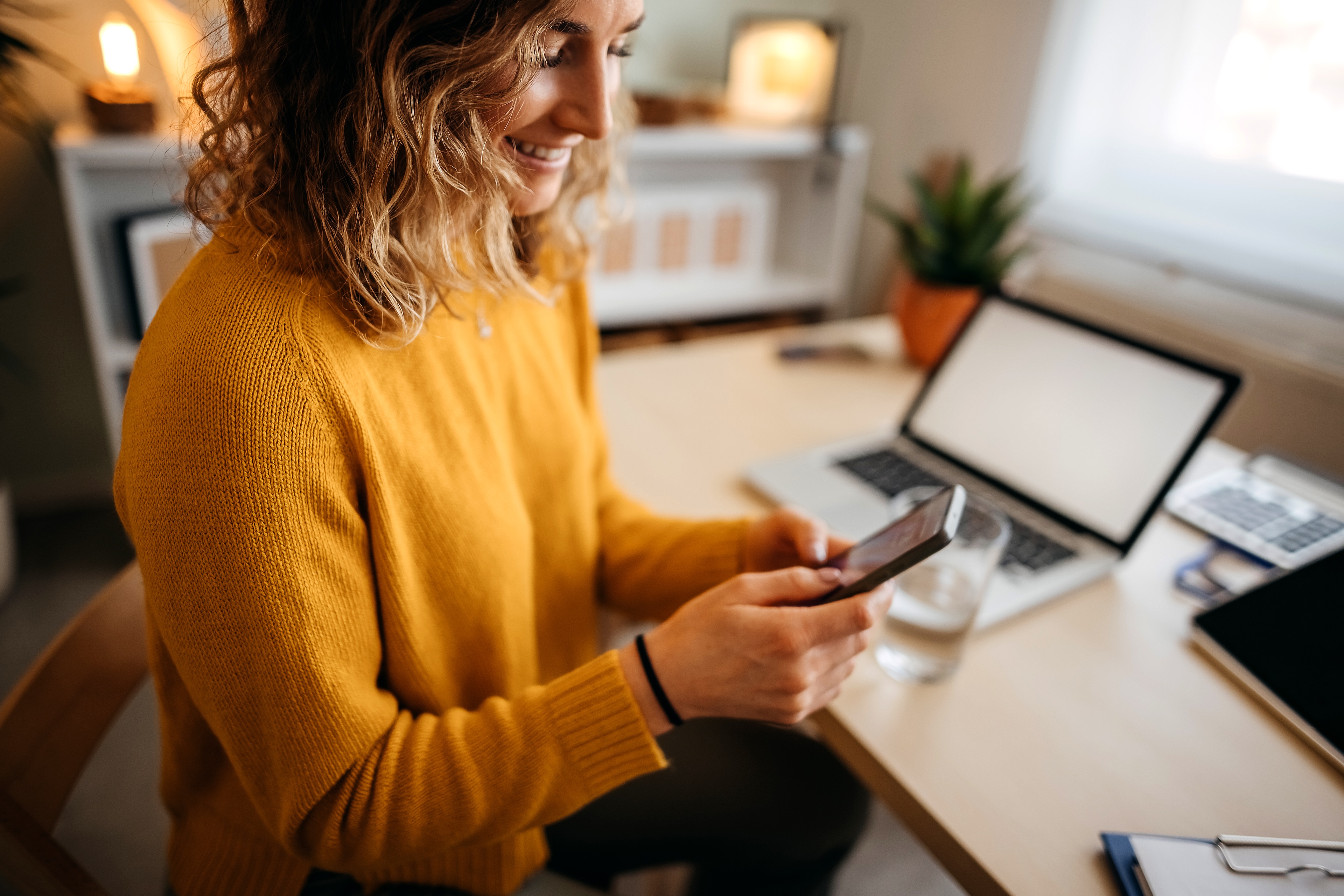 Frau mit orangenem Pullover steht in einem Büro und hält ein Smartphone in der Hand.