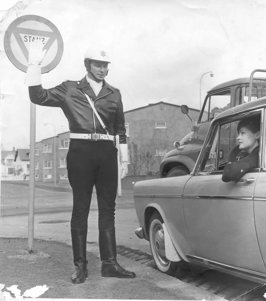 Lögreglumaður réttir upp aðra höndina fyrir ökumanni í bíl.