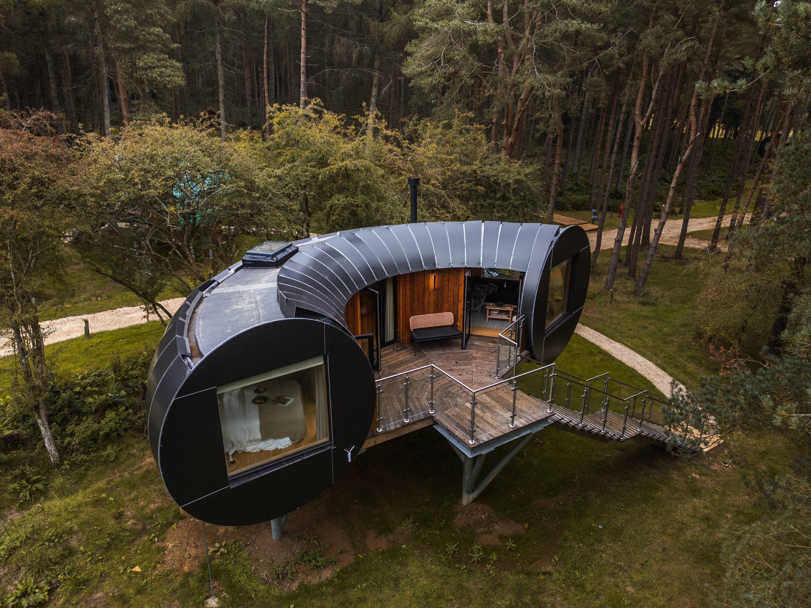 Soar above the enchanting Bastard Balm treehouse with this breath-taking aerial shot, capturing the seamless integration of modern architecture within the lush landscape. Nestled among the towering trees, this exterior view highlights the unique structure and design that make Bastard Balm a standout retreat in the Cotswolds. Experience the serene beauty of its natural surroundings, promising an unforgettable escape amidst the treetops.