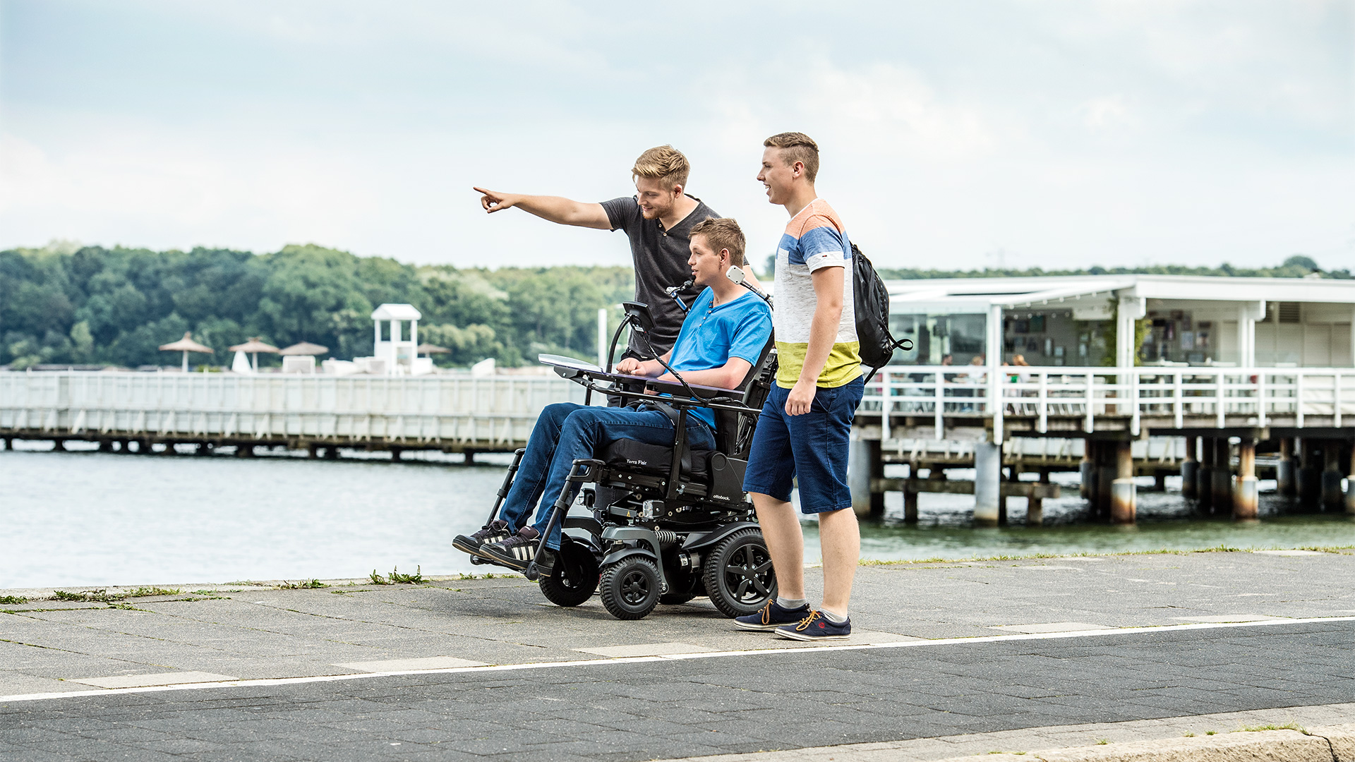 Three friends on a pier, one sitting in an Ottobock Juvo power wheelchair and another pointing at something in the distance