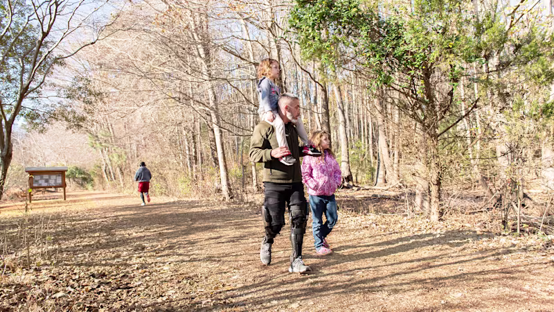 A family of three walking on an outdoor trail. The father holds his daughter on his shoulders without strain thanks to his custom-made Ottobock C-Brace