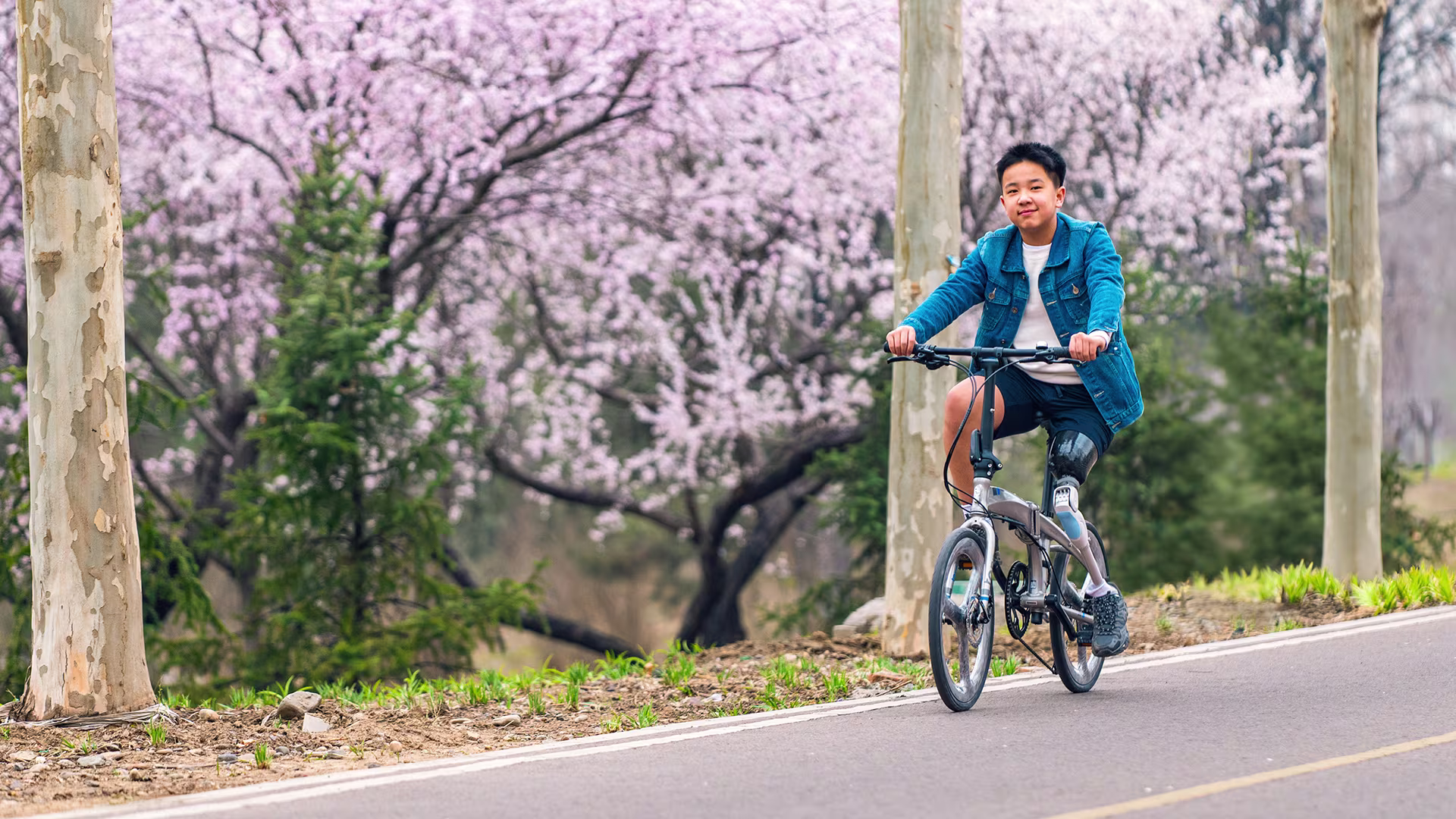 A teenage boy rides a bicycle in a park, wearing a prosthetic leg.