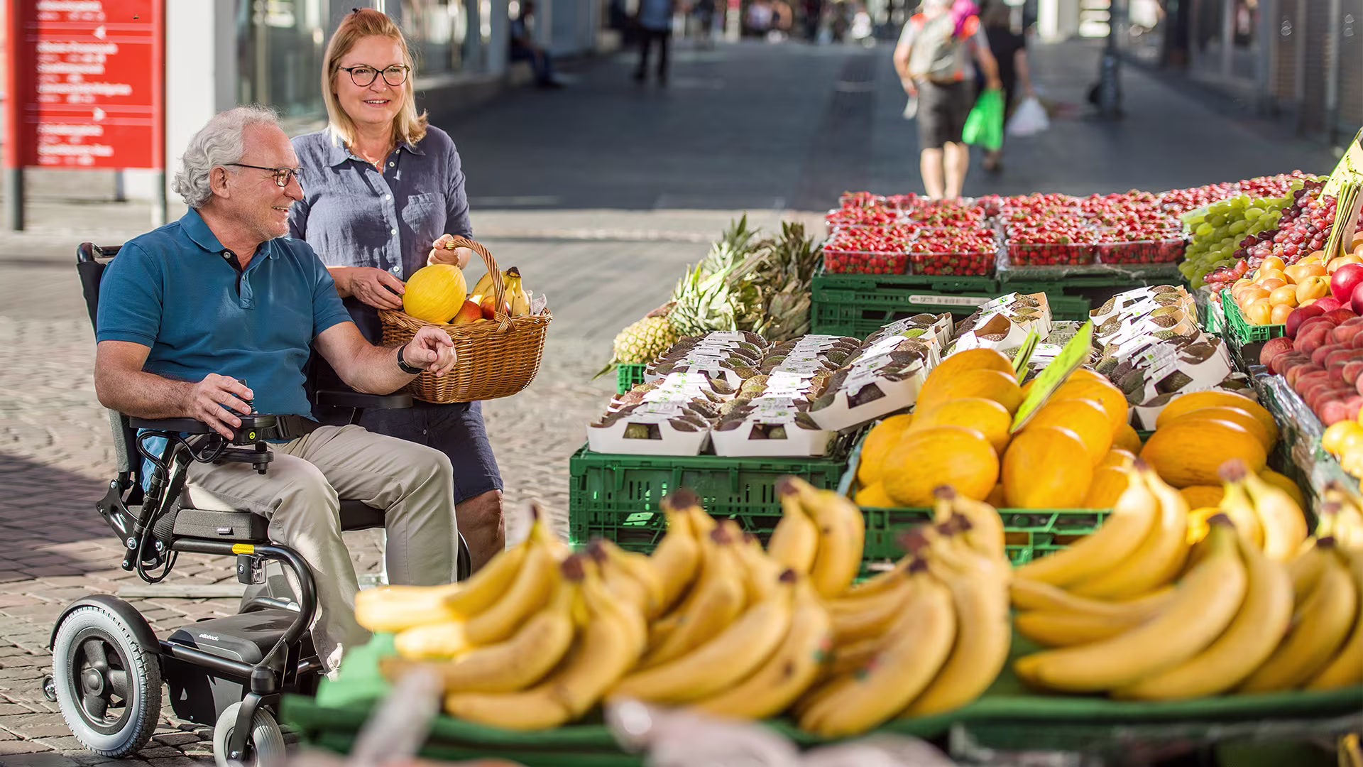 A man in an Ottobock wheelchair is smiling next to a woman with a shopping basket as they look at the products at a fruit market.