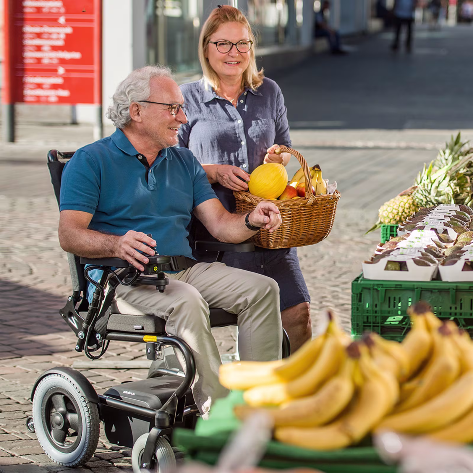 Auf dem Bild sieht man eine männliche Person in einem Rollstuhl sitzend und daneben eine weibliche Person stehend. Die beiden scheinen auf einem Markt Früchte einzukaufen.