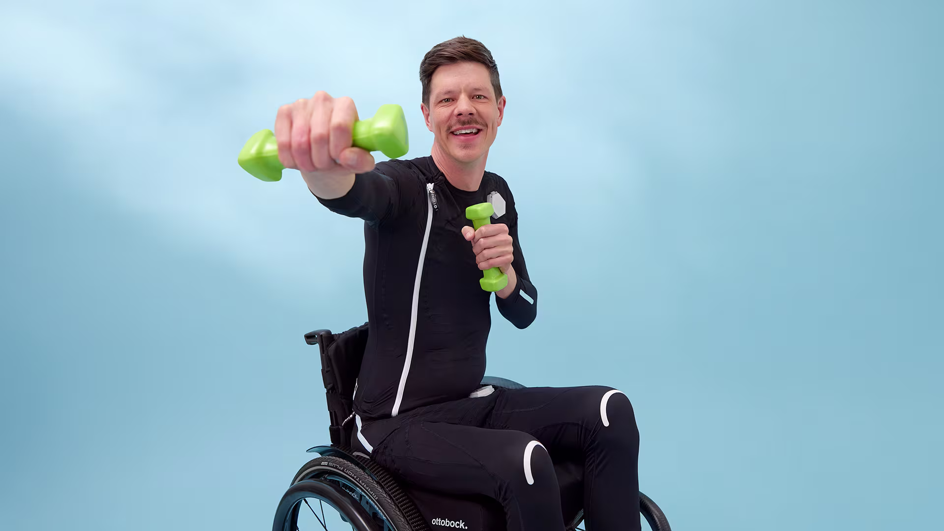 Ottobock Exopulse user Ricky sits in a wheelchair and smiles while holding two green dumbbells and stretching one Forward