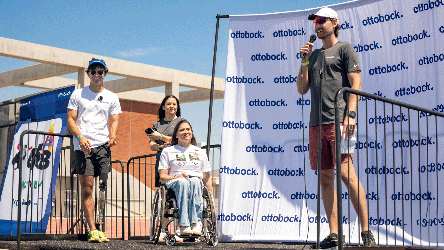 Clinical Specialist Lucas Crisanti on stage with a microphone in front of an Ottobock banner with three smiling onlookers  waiting their turn to speak