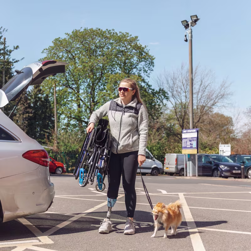 Hannah Moore holding her folded Avantgarde manual wheelchair under her arm whilst also holding her dogs lead.