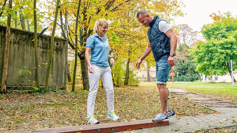 Amputee with Ottobock Kenevo MPK balances on a wooden beam with a smile while a prosthetist stands by his side