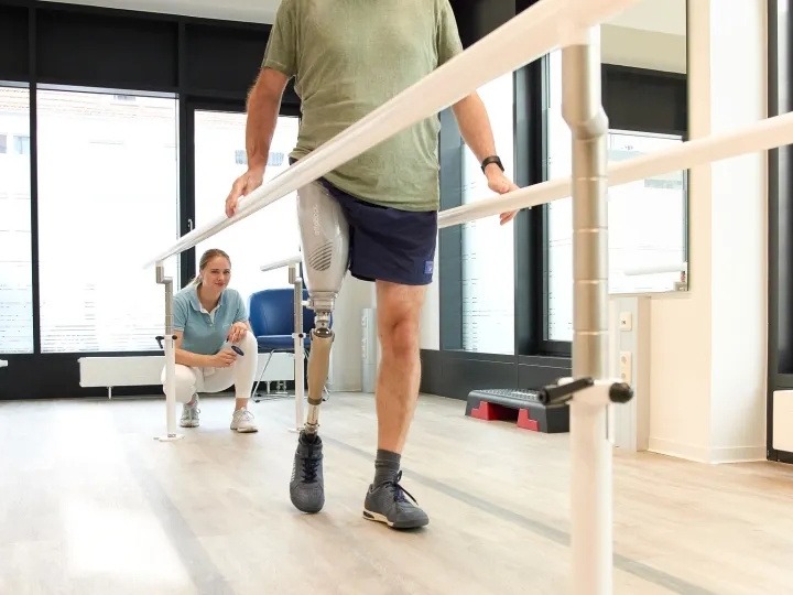 Man with above knee leg prosthesis walking between parallel bars during gait training, physiotherapist supervising in a rehabilitation clinic