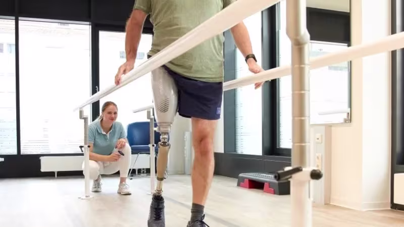 Man with above knee leg prosthesis walking between parallel bars during gait training, physiotherapist supervising in a rehabilitation clinic