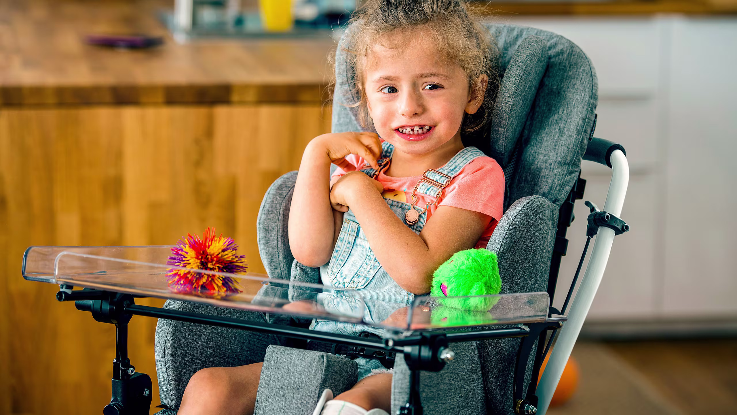 Bambino seduto nel seggiolone Ottobock con i giocattoli sul vassoio in cucina.