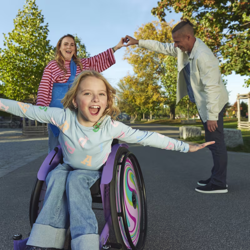 Nomine adlı genç bir kız, kidevo tekerlekli sandalyesinde kollarını yana uzatarak öne doğru eğiliyor, ebeveynleri arkasında dururken gülümsüyor, elleri bir kalp şekli oluşturacak şekilde birleşiyor.