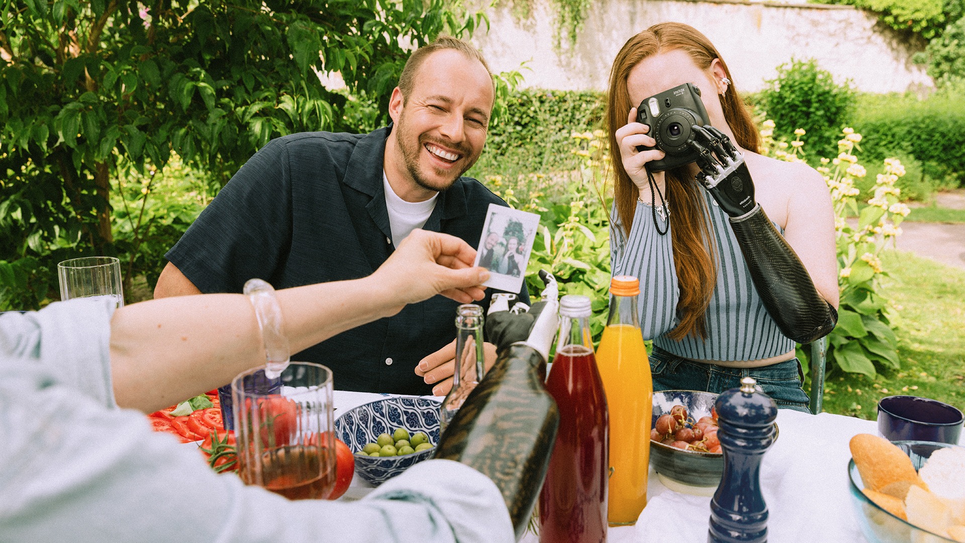 A man and a woman with prosthetic arms and a camera in her hand sit at a table set for summer.