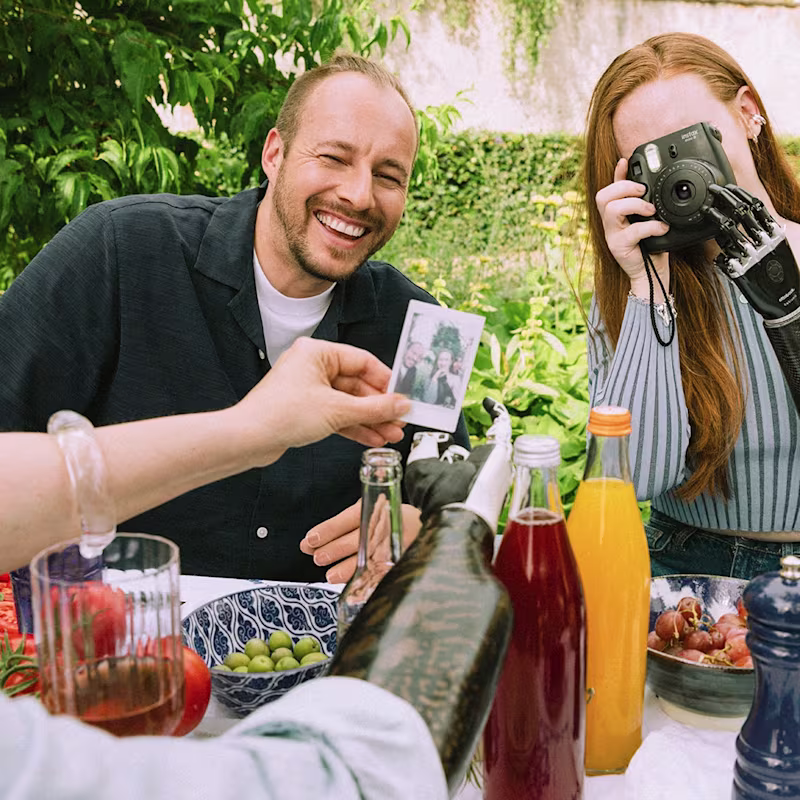 A man and a woman with prosthetic arms and a camera in her hand sit at a table set for summer.