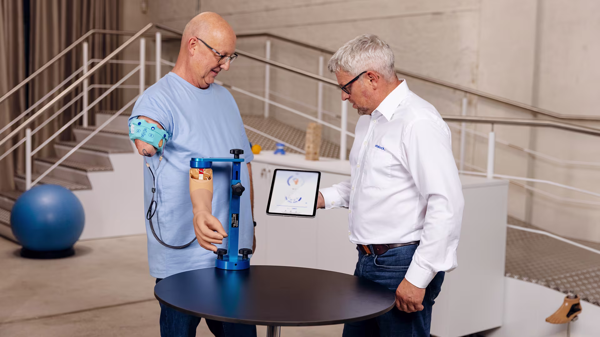 Ralf, wearing a myoelectric prosthesis, sitting with a technician who is assisting him in adjusting MyoBock prosthetic settings using the connectgrip app on a tablet.