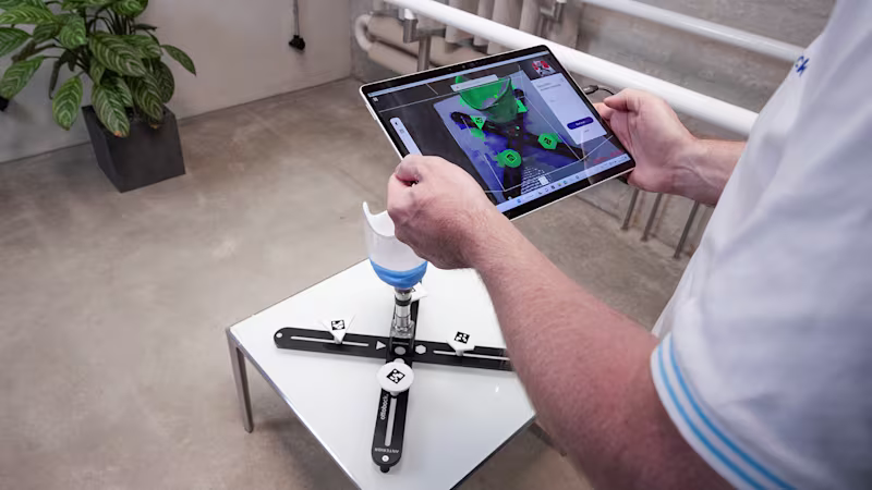 Person nutzt ein Tablet, um mit einem Prothesen-Anpassungssystem zu interagieren. Auf dem Bildschirm werden digitale Markierungen auf einer Prothesenkomponente angezeigt, die auf einem Kalibrierungsständer in einem modernen, minimalistisch eingerichteten Arbeitsbereich positioniert ist.
