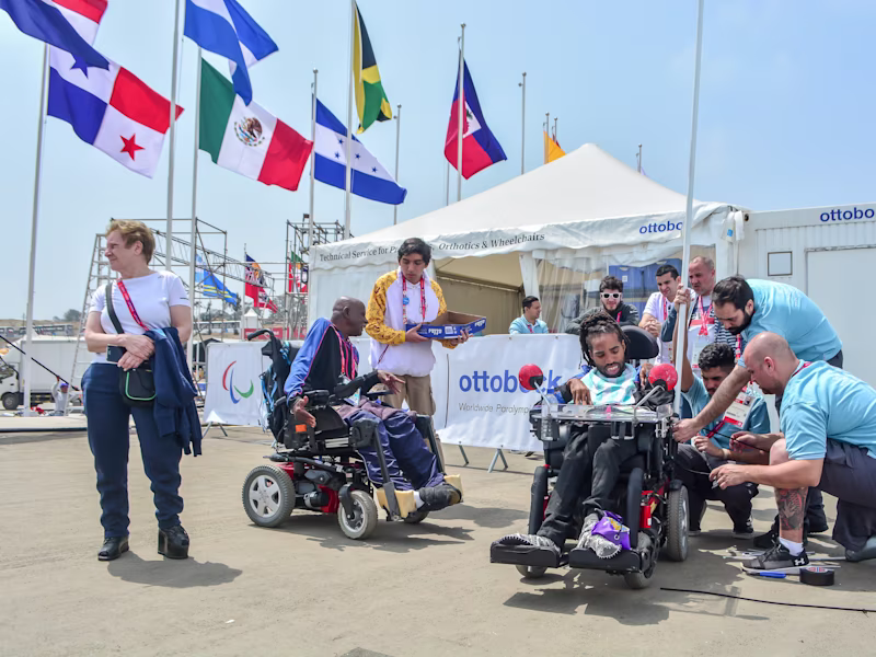 Ein Team von Ottobock-TechnikerInnen repariert den Rollstuhl einer männlich gelesene Person. Im Hintergrund sind Flaggen und die Kulisse der Paralympischen Spiele zu sehen.