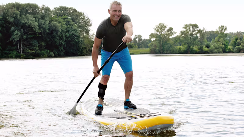 Eine männlich gelesene Person paddelt auf einem Stand-Up Paddle Board auf dem See und trägt dabei eine Orthese als Hilfsmittel.