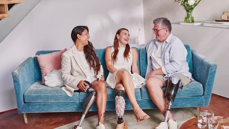 Three lower limb prosthetic users with transtibial and transfemoral prostheses sit on a sofa, talk and laugh.