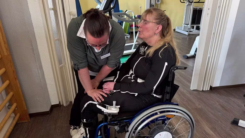 Auf dem Bild sieht man Diana, die in ihrem Rollstuhl sitzt und ihren Exopulse Mollii Suit Anzug trägt. Eine andere Frau steht vor ihr gebeugt und bewegt Dianas Knie.