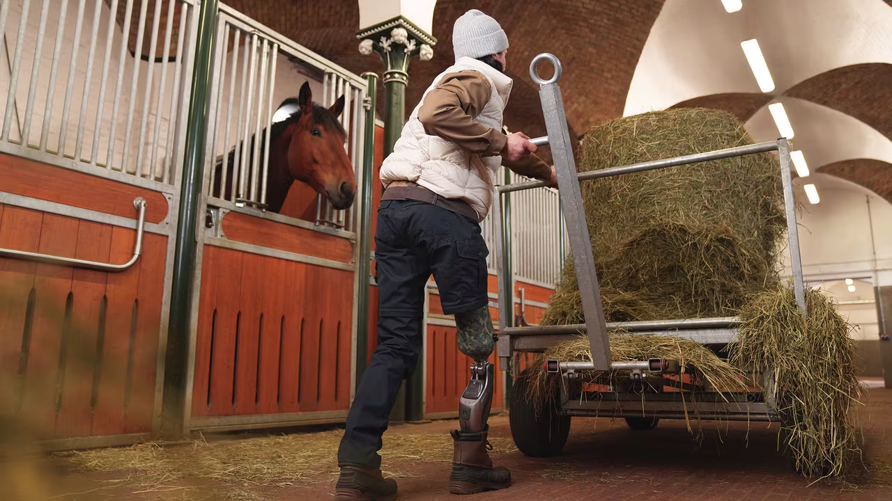 Eine Prothesenträgerin schiebt einen Wagen mit Stroh durch einen Pferdestall, um Ihr Pferd zu füttern. Sie trägt das mechatronische Prothesenkniegelenk Genium X4 von Ottobock.