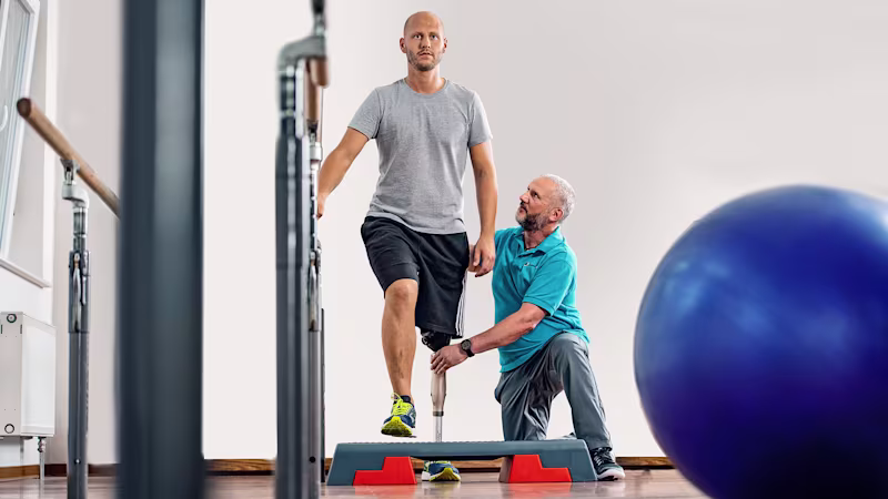 Ein oberschenkelamputierter Mann stützt sich an einem Barren ab und steigt auf ein Step-Brett. Er trägt dabei das mechatronische Kniegelenk Kenevo von Ottobock und wird von einem Therapeuten unterstützt.