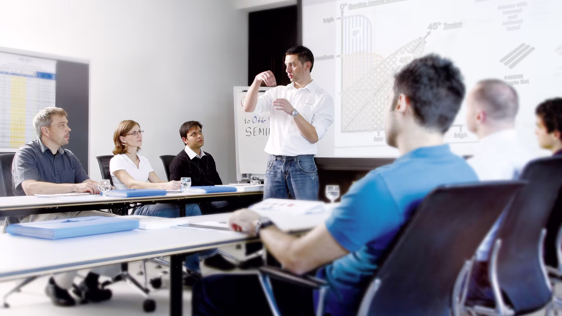 Eine männlich gelesene Person steht vor einigen TeilnehmerInnen und hält einen Seminarvortrag.