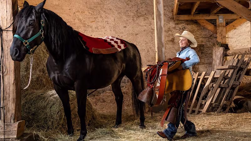 A person with a transtibial amputation carries a saddle toward a horse.
